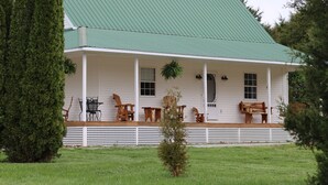 A white house with a green roof, a porch with wooden furniture, and a small tree in the front yard. - Good Hope Cottage- Amish Country / fire pit (Lawrenceburg)