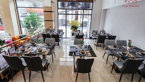 A hotel dining area with multiple tables and chairs, large windows with a view of the street, and a logo on the wall. - Ciao SaiGon 2 Hotel (Ho Chi Minh City)