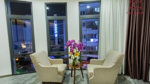 A corner room with two beige armchairs, a small wooden table with a vase of flowers, and large windows with city views. - Ciao SaiGon 2 Hotel (Ho Chi Minh City)