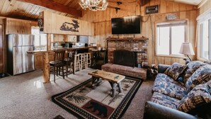 Interior rústico de una cabaña con comedor de madera, chimenea, sofá y televisor en la pared.