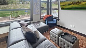A modern living room with a large gray sectional sofa, a blue armchair, and a small ottoman. A large window offers a view of a green landscape with houses and trees. - Best Location in Snowmass Village! With A/C, Amazing Views, Balcony, W/D, Gas FP & Shared HT (Snowmass Village)