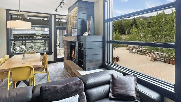 A modern living room with a fireplace, large windows, a dining area, and a view of a park. - Best Location in Snowmass Village! With A/C, Amazing Views, Balcony, W/D, Gas FP & Shared HT (Snowmass Village)