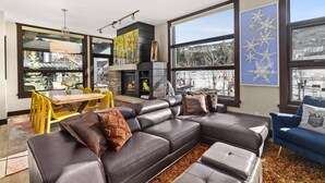 A modern living room with a sectional sofa, a dining table with chairs, a fireplace, and large windows offering a view of the outdoors. - Best Location in Snowmass Village! With A/C, Amazing Views, Balcony, W/D, Gas FP & Shared HT (Snowmass Village)