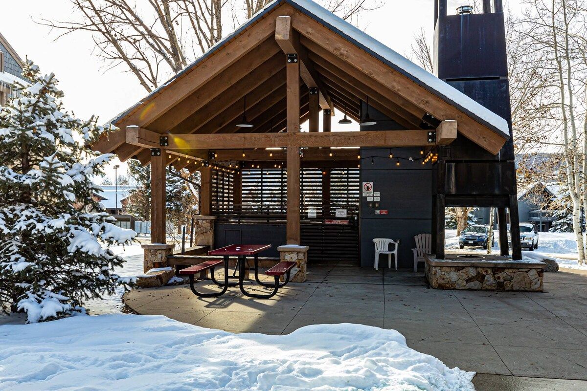 A covered outdoor area with a wooden structure, picnic tables, and a stone bench.