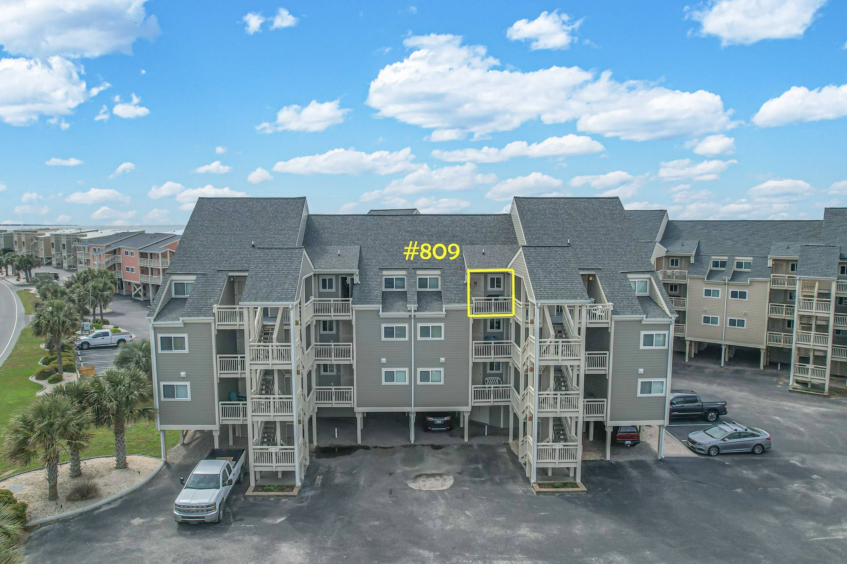 A multi-story residential building with a grey facade, balconies, and a parking area.