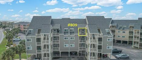 A multi-story residential building with a grey facade, balconies, and a parking area.