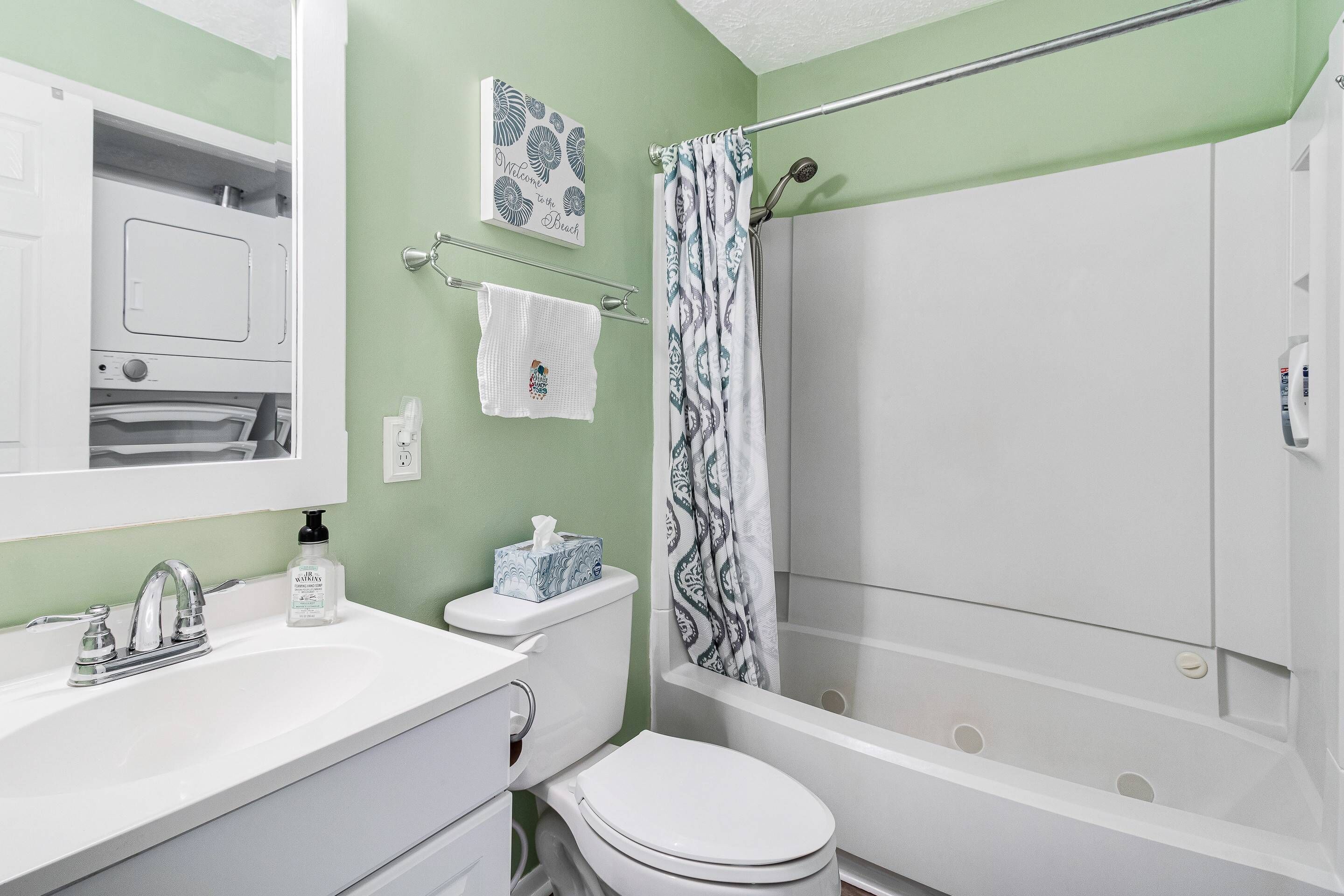 Ein Badezimmer mit einem weißen Waschbecken, einer Toilette und einer Dusche. Ein Handtuchhalter mit einem weißen Handtuch sowie ein blau-weiß gemustertes Duschvorhang sind vorhanden. Ein Willkommensschild befindet sich an der Wand.