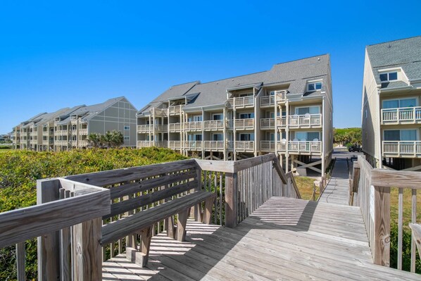 A wooden boardwalk leading to a beachfront hotel with multiple balconies and a clear view of the ocean.