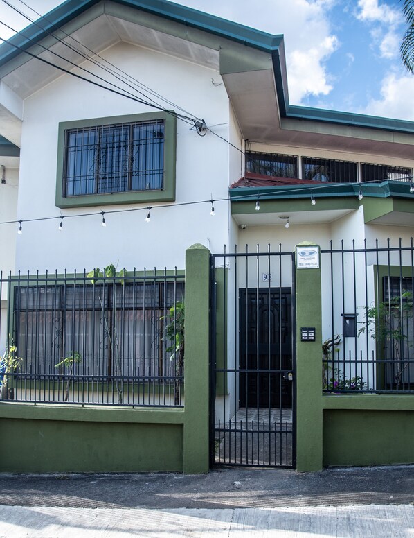 Un edificio de dos pisos con fachada verde y blanca, entrada con portón y un pequeño balcón con techo rojo.