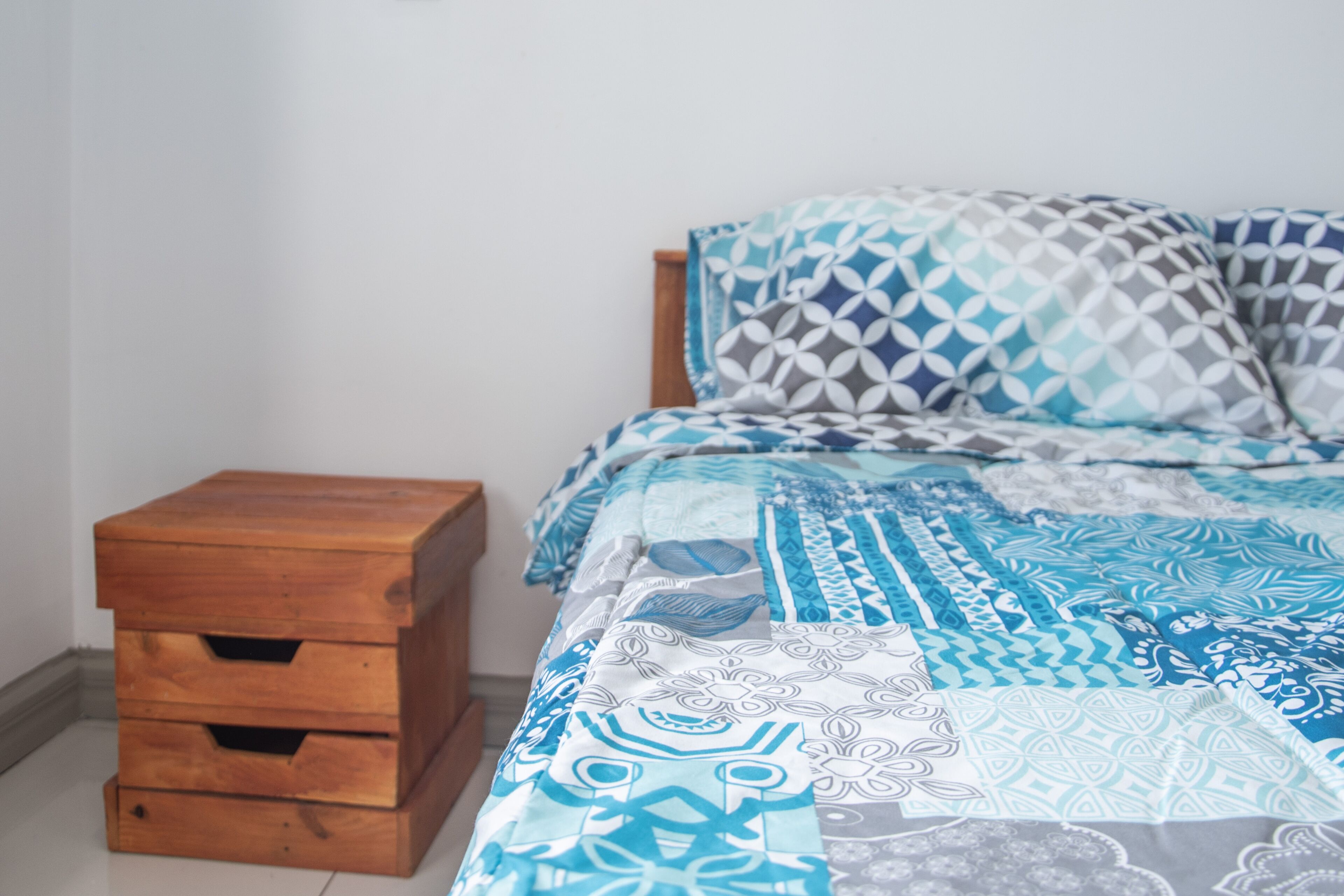 Une chambre à coucher avec un lit recouvert d’une literie à motifs, une table de chevet en bois dotée de tiroirs et un mur uni.
