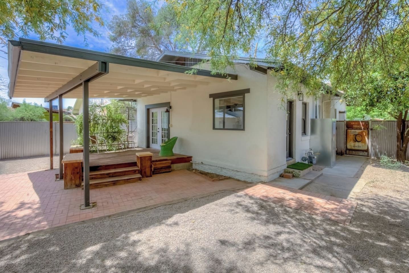 A covered patio area with a wooden bench and a green chair, a small outdoor seating area, and a white building with a door and windows.