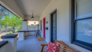 A covered patio with a bench and a table, a red door, and a window with a view of greenery.