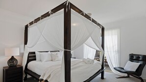 A four-poster canopy bed with white linens, a dark wooden frame, and a black leather chair in a modern room with a neutral color palette. - Renovated open planned living sun drenched house in Cape Woolamai (Cape Woolamai)