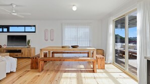 A spacious living room with a wooden floor, a large TV, a dining table with benches, and a view of a deck through large windows. - Renovated open planned living sun drenched house in Cape Woolamai (Cape Woolamai)