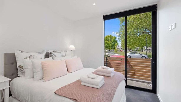 A modern bedroom with a large bed, a nightstand, a window with a view of the street, and a sliding door. - Latimer View Apartment (Christchurch)