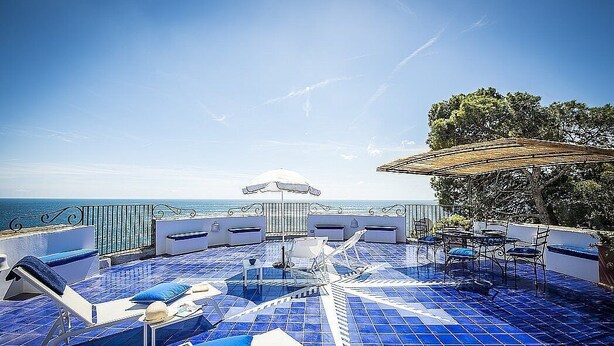 A rooftop terrace with blue-tiled flooring, lounge chairs, and a table with umbrellas.