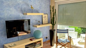 Un salon avec un téléviseur, des étagères, une plante et un fauteuil près d’une porte-fenêtre coulissante.