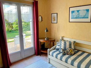 A room with a bed, a table, a lamp, a window with curtains, and a framed picture on the wall.