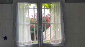 Une fenêtre avec des rideaux blancs, d’où l’on aperçoit des fleurs roses à l’extérieur.