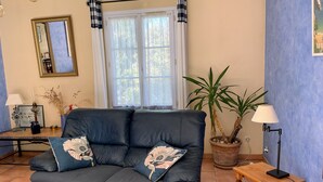 Un salon avec un canapé en cuir foncé, une table d’appoint en bois, une plante en pot, un miroir et une fenêtre avec des rideaux.