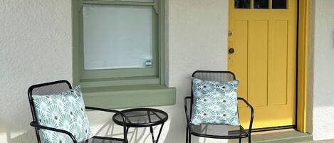 A porch with two chairs and a table, a yellow door, and a window with a green frame.