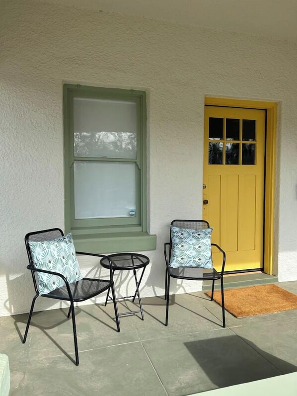 A porch with two chairs and a table, a yellow door, and a window with a green frame.