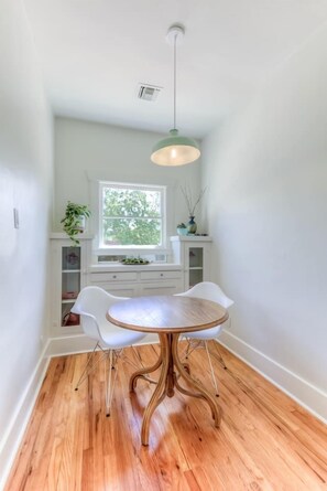 A dining area with a round wooden table, white chairs, a hanging light fixture, and a window with a view of greenery. - Remodeled Gem 5 minutes to UA (Tucson)