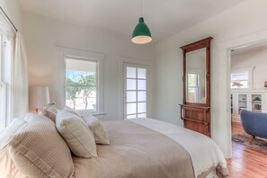 A bedroom with a large bed, a mirror, a dresser, and a chair. There are windows and a door.