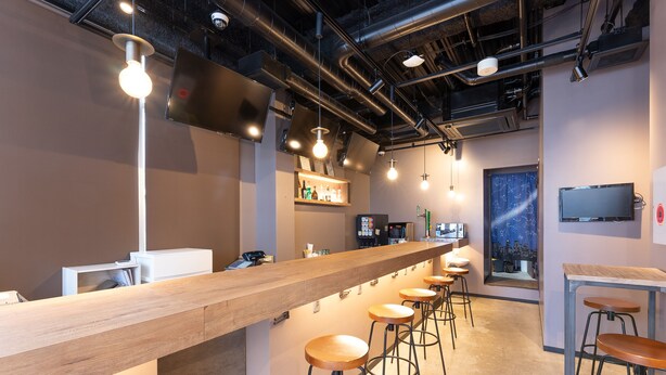 A modern bar area with a long counter, multiple stools, and overhead lighting. - Tabist MAYUDAMA CABIN (Yokohama)