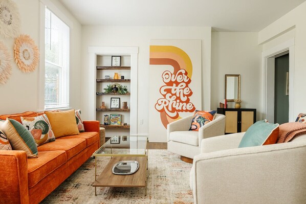 Uma sala de estar com um sofá laranja, uma poltrona branca e uma mesa de centro. Há uma estante com objetos decorativos e uma grande obra de arte na parede com a inscrição 'Over the Phine'.