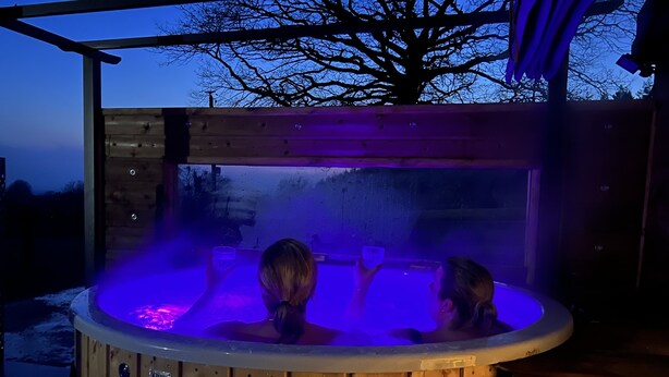 Un bain à remous en bois, doté d’une enceinte en verre, entouré d’une terrasse en bois et surmonté d’une structure de toit en métal.