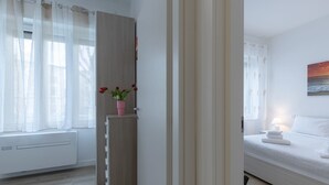 Un dormitorio con cama, ventana con cortinas, radiador, armario y un florero con flores.