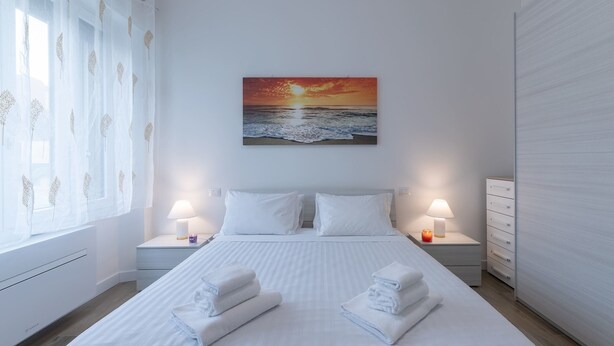Habitación de hotel con cama, dos mesitas de noche, una lámpara y un cuadro de un atardecer sobre el océano.