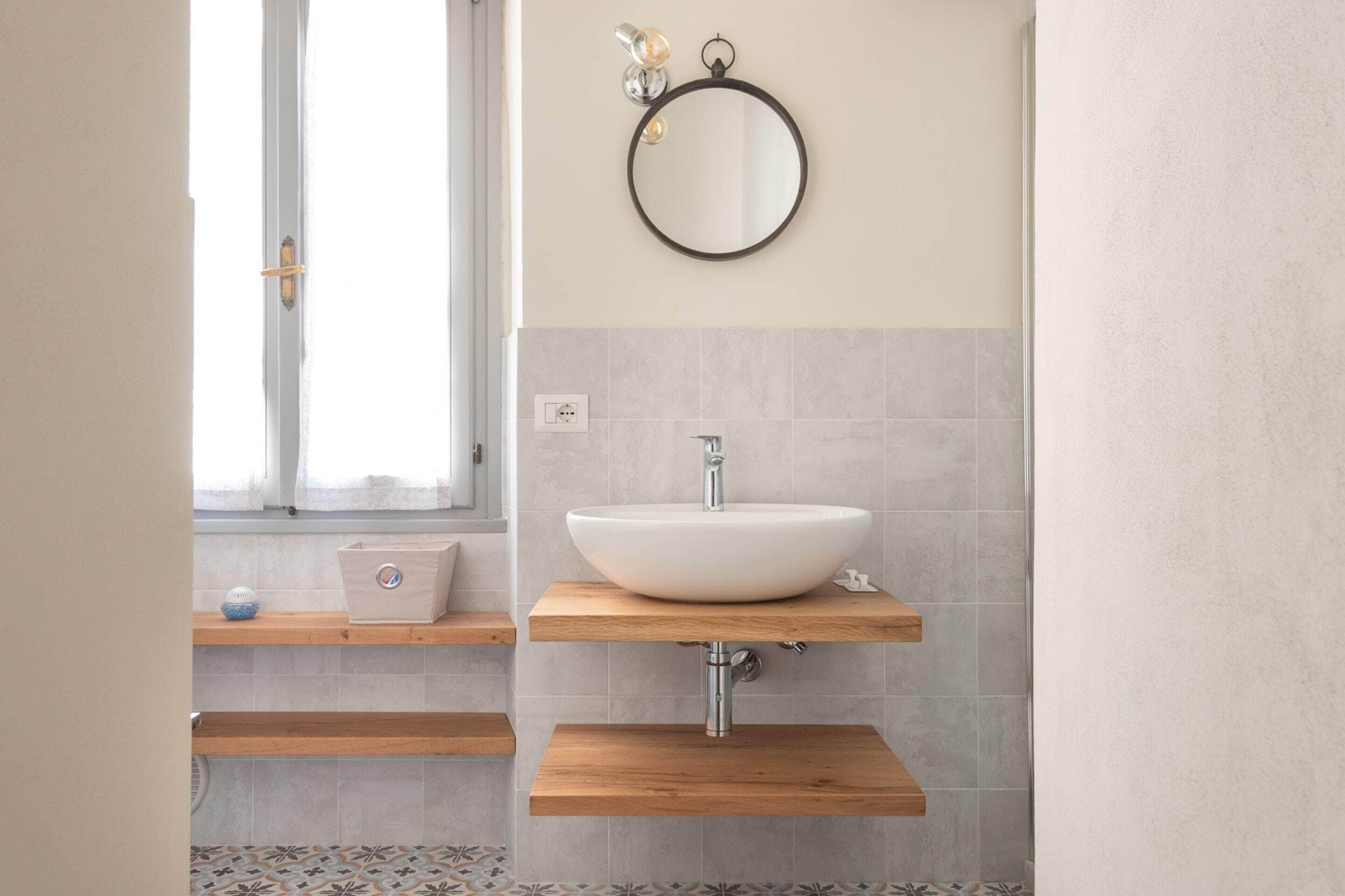 A bathroom with a white sink, wooden shelves, a round mirror, and a window with a view.