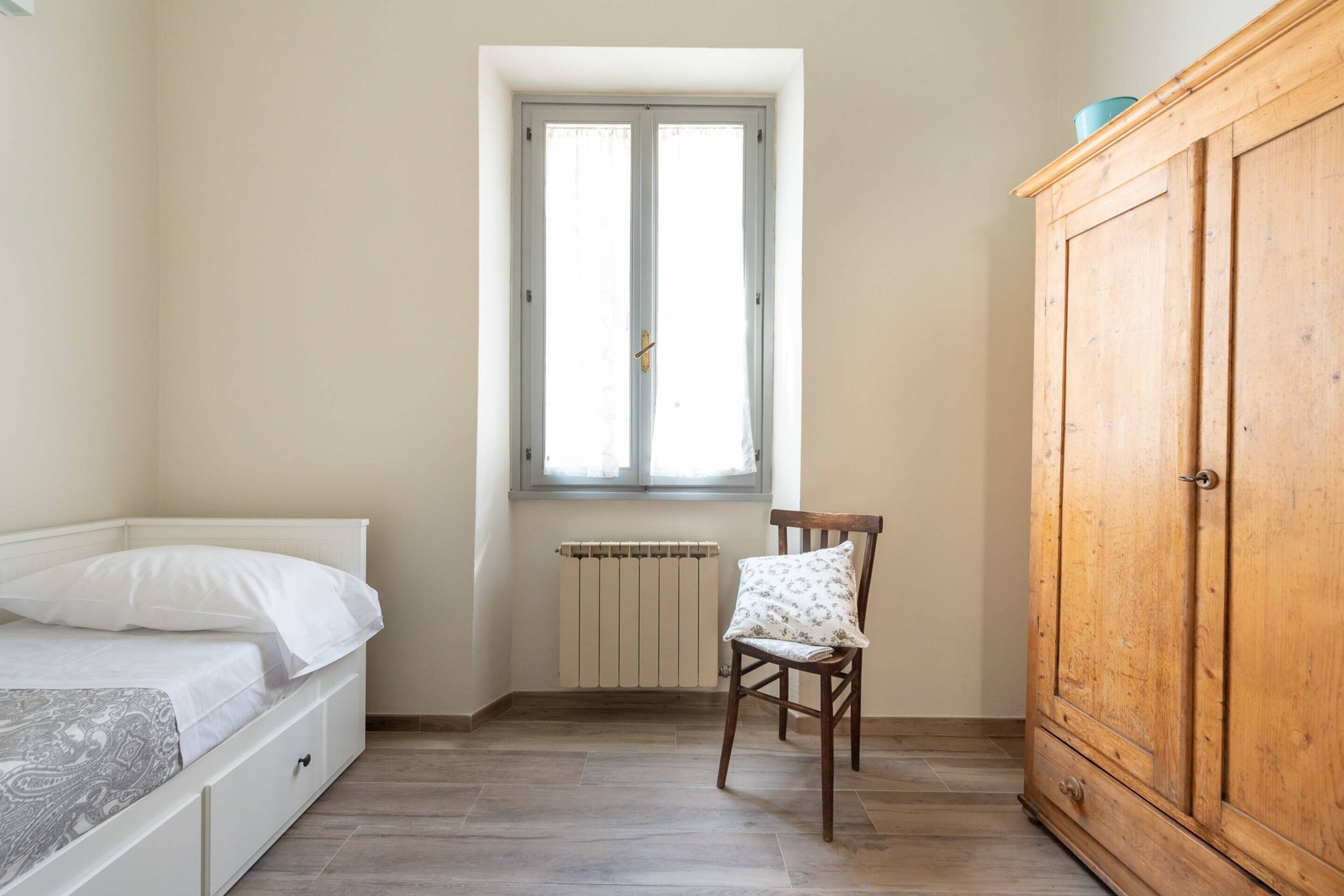 Et soveværelse med seng, radiator, et vindue, en stol og et træskab.