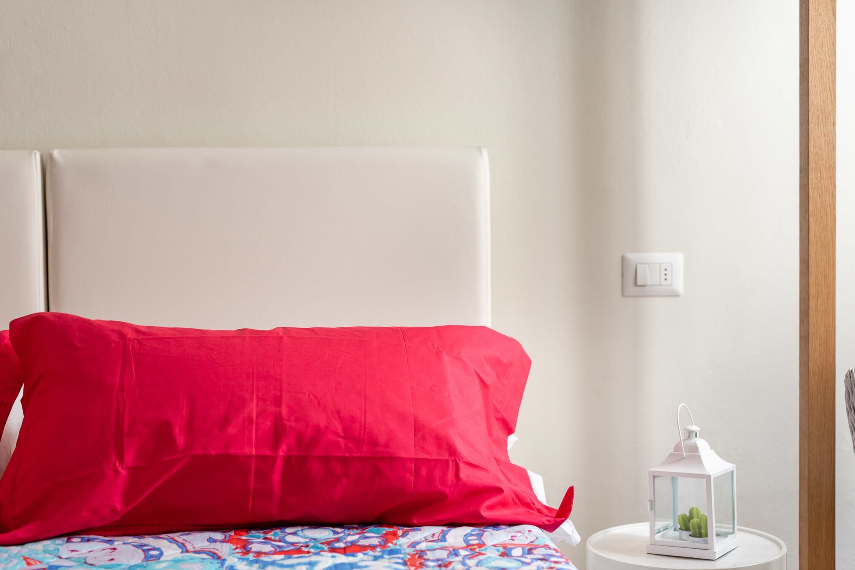 A bed with a red pillow and a patterned bedspread, a white nightstand with a white lantern, and a white wall with a light switch.