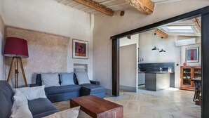 Uma sala de estar moderna com um sofá, mesa de centro e abajur. Uma cozinha fica visível por uma porta aberta.
