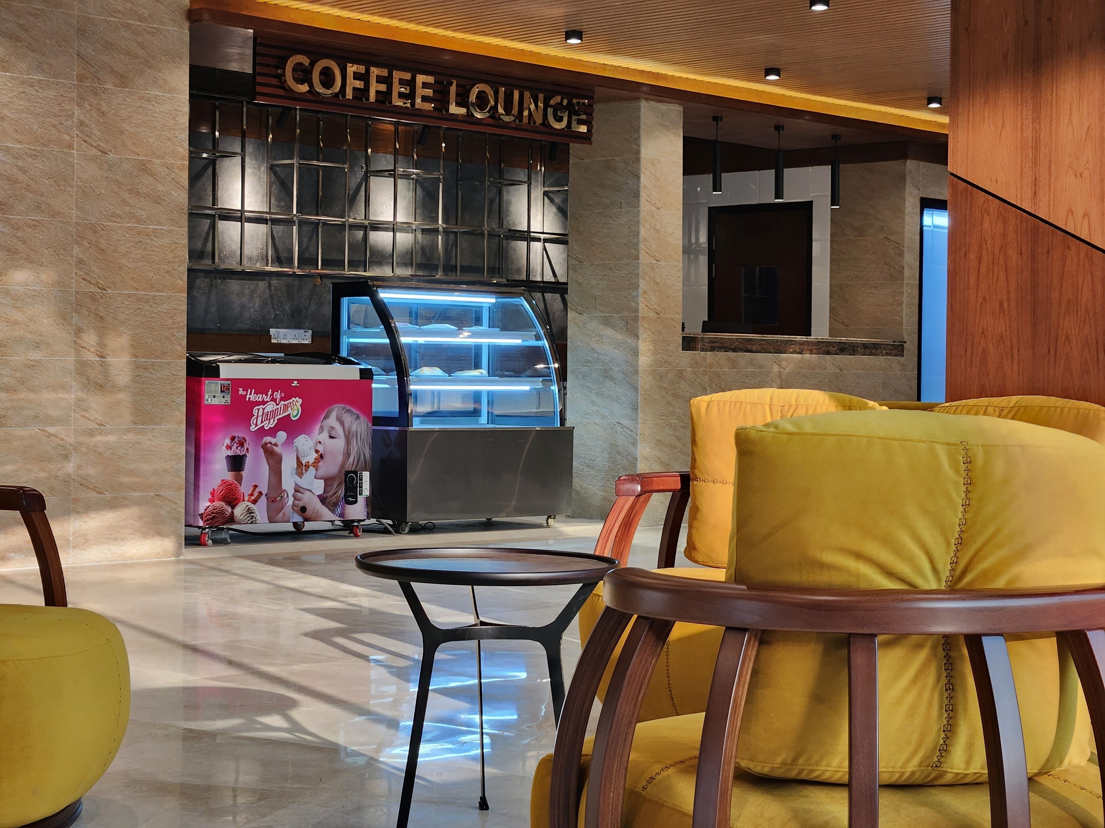 Un espace détente avec un coin café au design moderne, comprenant un comptoir avec une vitrine et une affiche promotionnelle. On y trouve des chaises jaunes et une petite table au premier plan.