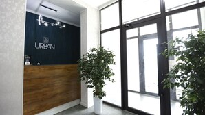 A reception area with a wooden desk, a dark blue wall with the word "URBAN" on it, and a large window with a view outside. - Urban Hotel (Bishkek)