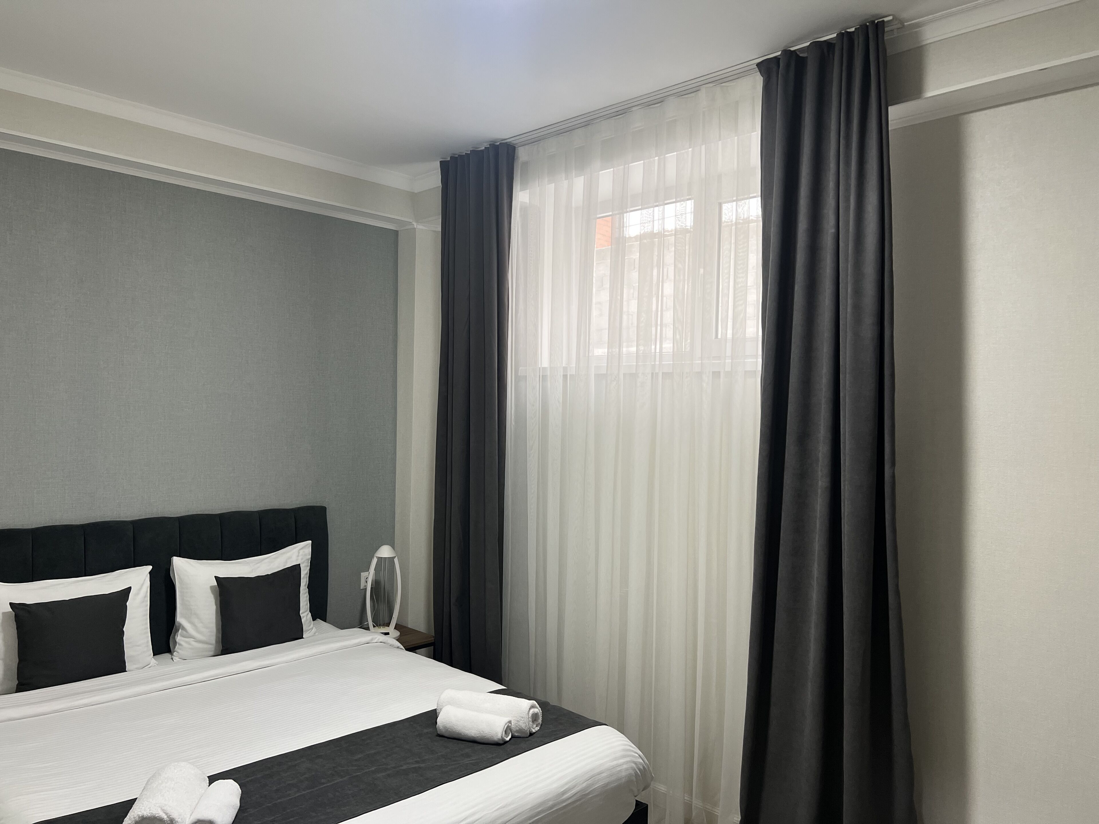 A hotel room with a neatly made bed, a bedside lamp, and sheer curtains.