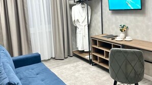 A modern hotel room with a blue sofa, a grey armchair, a wooden desk with a flat-screen TV, a clothing rack, and a large window with curtains. - Urban Hotel (Bishkek)