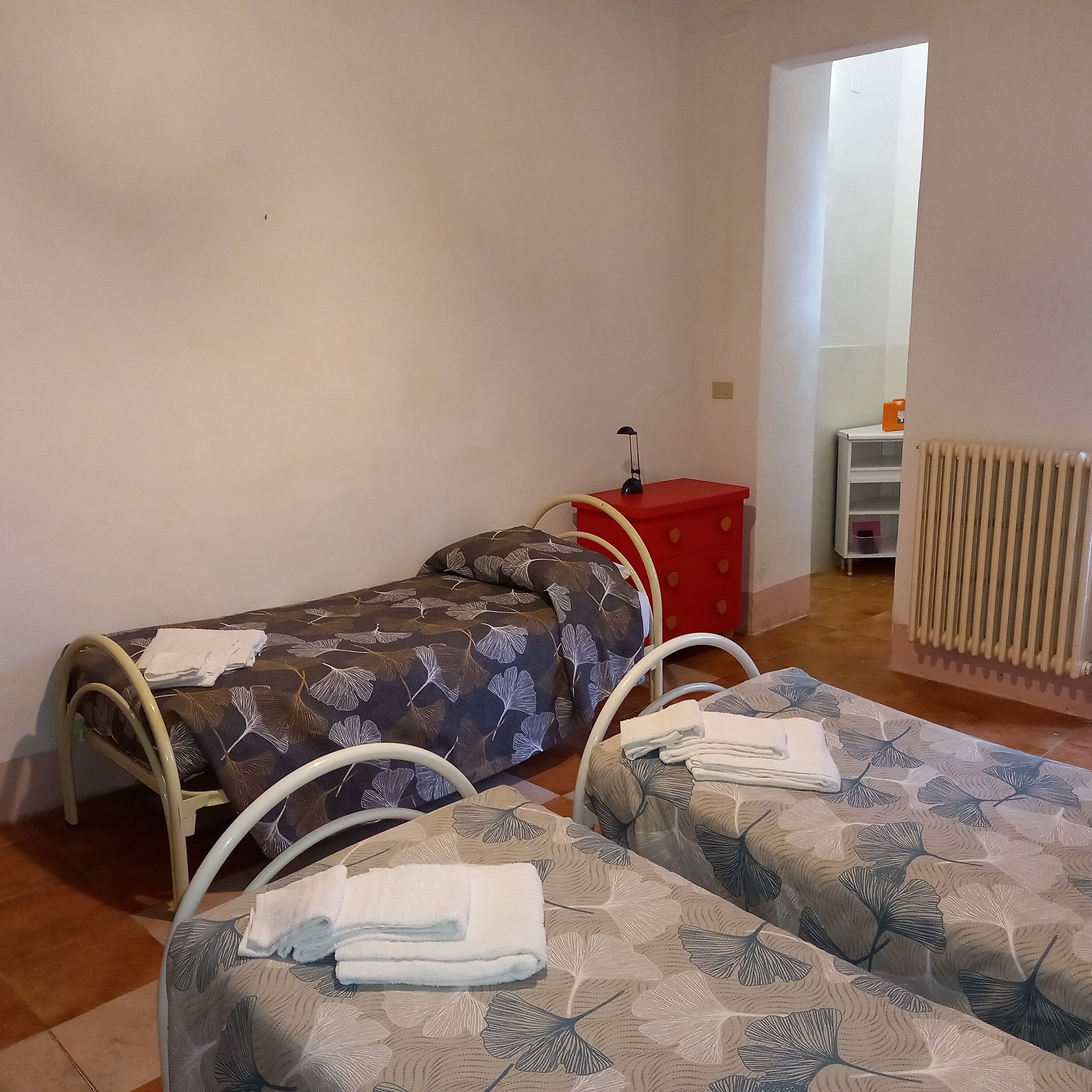 A small, tidy room with a single bed, a red nightstand, a white radiator, and a window.