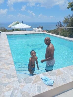 A swimming pool with a tiled edge, a white umbrella, and a view of the ocean.