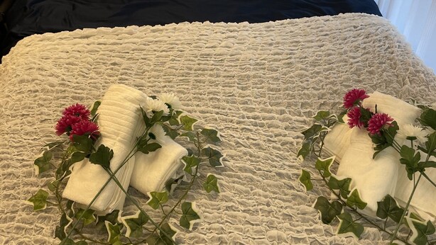 Siististi pedattu sänky, jossa on valkoiset ja tummansiniset vuodevaatteet, kaksi kukkapainokuosisia pyyhettä ja kuvioitu valkoinen peitto.