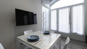Uma área de refeições moderna com televisão de tela plana na parede, uma mesa de jantar com pratos e um copo, e vista para uma janela grande com cortinas.