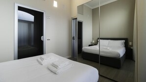 Quarto de hotel moderno com uma cama grande, uma mesinha de cabeceira com abajur e um espelho refletindo o ambiente.