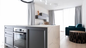 Une cuisine moderne dotée d’un îlot en marbre, d’un four encastré et de meubles de rangement gris. On y trouve également un fauteuil turquoise et une table ronde en bois au design original.