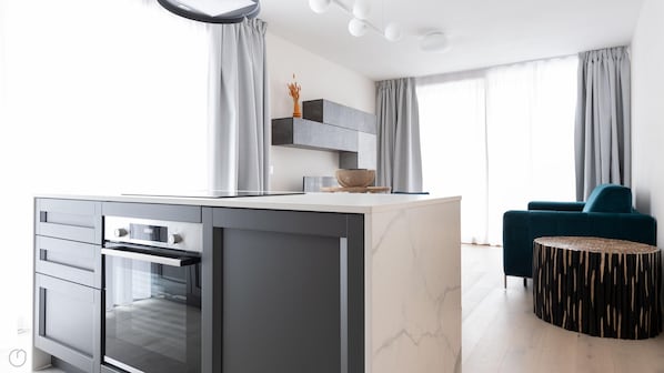 A modern kitchen with a marble island, built-in oven, and grey cabinetry. There is a teal armchair and a round wooden table with a unique design.