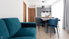 A modern living room with a teal sofa, a dining area with a table and chairs, and a kitchen with grey cabinets and a hanging light fixture.