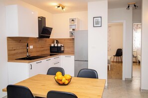 A modern kitchen with white cabinets, a wooden countertop, a black stove, and a stainless steel refrigerator. There is a dining table with a fruit bowl and black chairs.